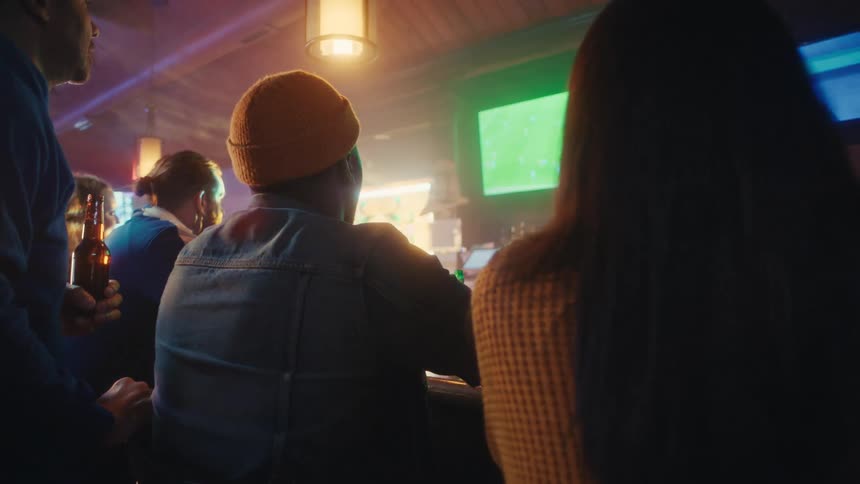 Small group of people watching a soccer game on the the television at the counter of a bar while the bartender is serving on the background indoors during night time