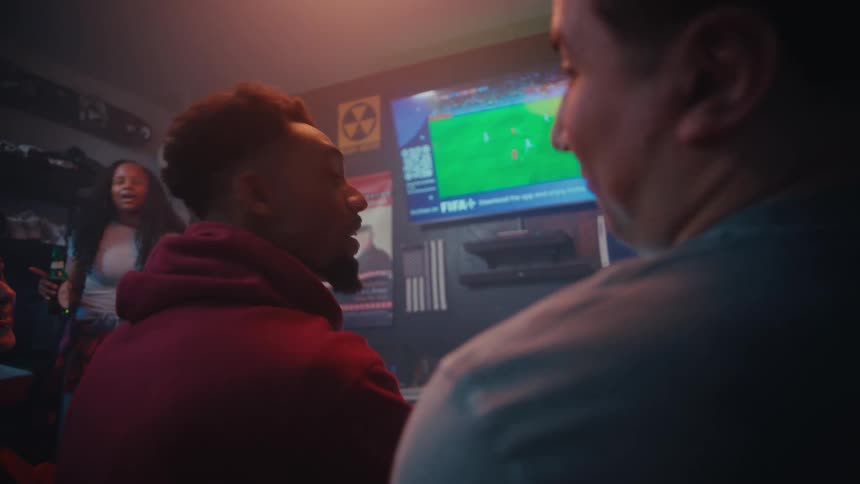 Two adult men talking while watching a soccer game at a pub with other people around them indoors during night time