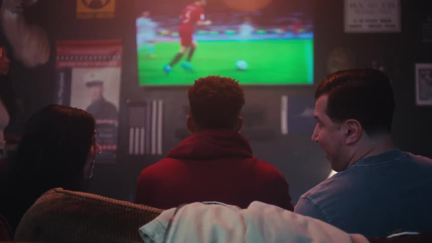 Three people are sitting on a couch as one watches a soccer match and the other two talk