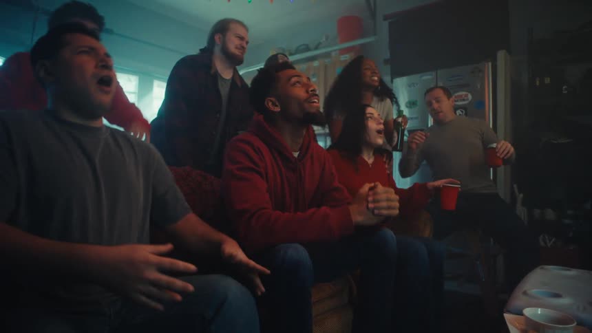 A group of friends watches a screen with intense focus and anticipation while gathered in a garage their expressions suddenly shift to collective disappointment and frustration as they react to a negative turn of events