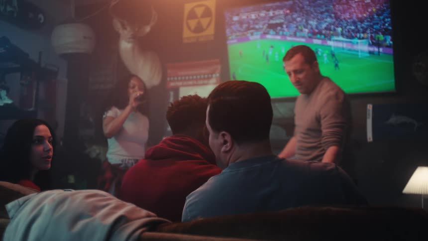 A group of friends sits in a dimly lit room focusing intently on a soccer match playing on a large television screenwhile some stay seated on the couch others move around the room with drinks occasionally glancing at the game or talking to one anot