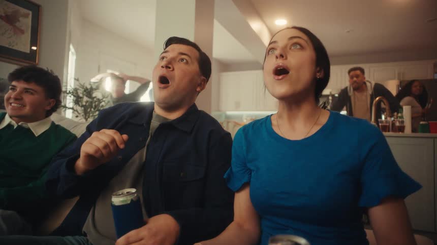 People sitting on the couch in front of the television and standing in the kitchen at a house party when they get excited and cheer