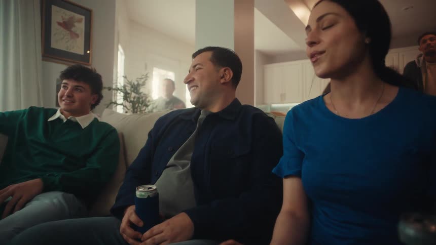 A group of friends sitting in a living room holding beer cans and watching a game with excitement