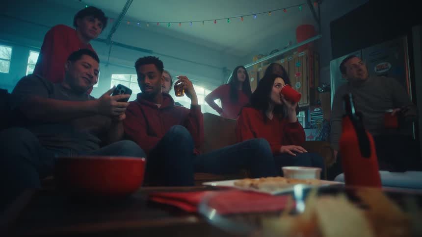 A group of friends sits in a dimly lit garage decorated with string lights focused on a screen while sharing drinks and snacksas the scene progresses the man on the left pulls out his phone to show something to the group sparking a collective react