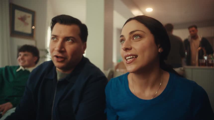 A man and a woman sit closely together indoors their faces lighting up with excitement as they watch something offcamera their expressive reactions and wide smiles suggest they are sharing a lively and engaging moment with others in the background
