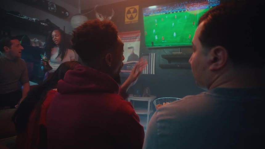 A group of friends watches a soccer match on television while talking and eating snacks in a dimly lit room