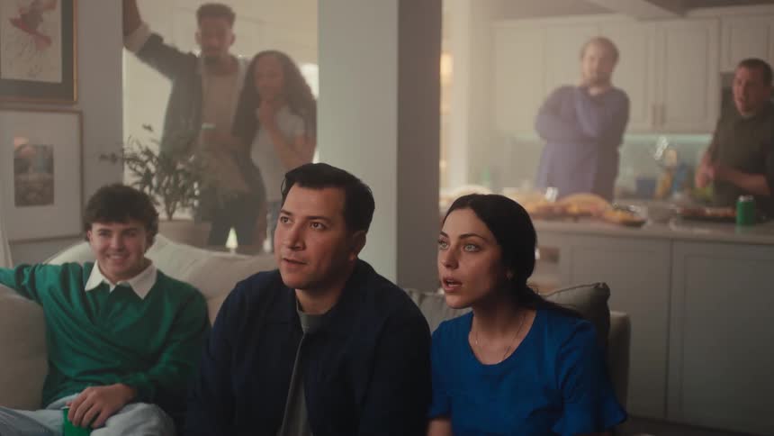 Group of people seated in the living room chat while others prepare food in the kitchen in the background