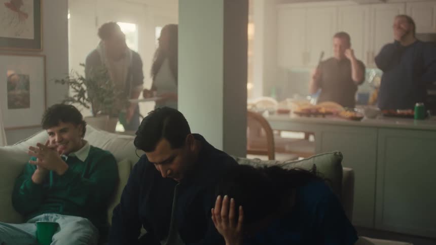 People seated on a couch reacting to something offscreen while others move around the kitchen in the background during a social gathering