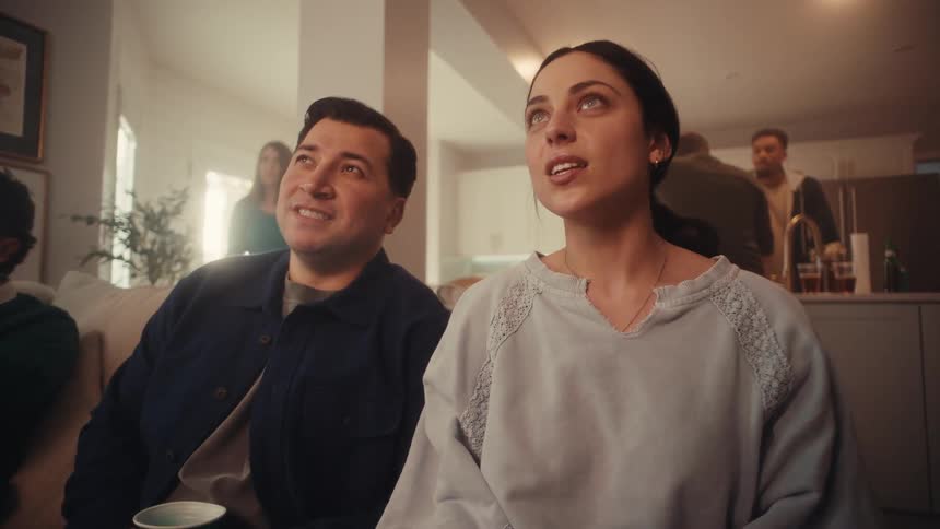 Group of people seated in a living room watch an offscreen event while others move in the background