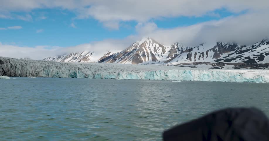 A boat moving toward an iceberg near the shoreline with a snowcovered mountain range in the background at midday