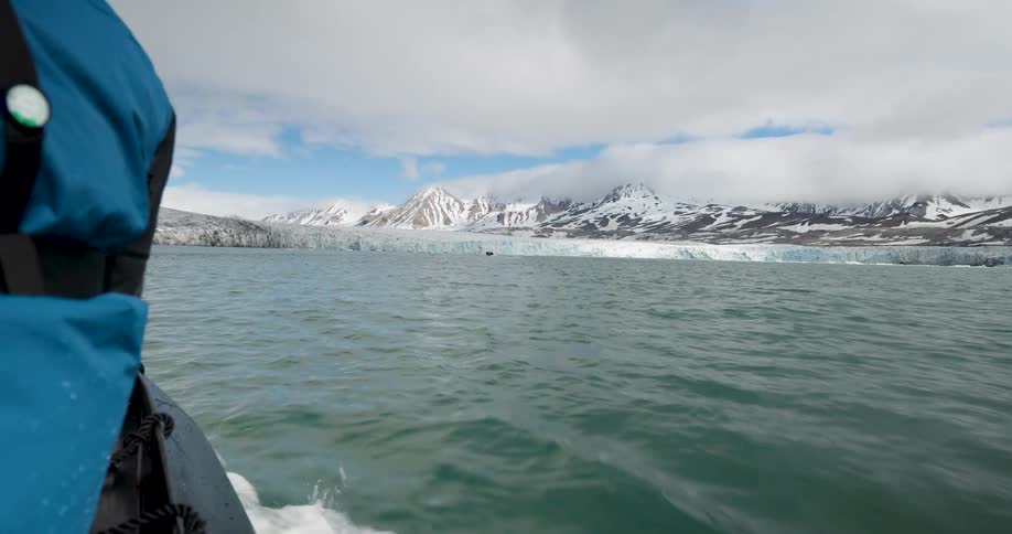 A boat travels through choppy arctic waters toward a massive bright blue glacier front nestled at the base of snowcovered mountainsclouds hang low over the rugged peaks while the wake of the vessel ripples through the dark sea in the foreground