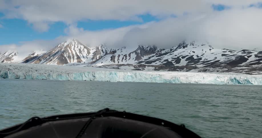 A boat moving across a lake toward a frozen shoreline with a snowcovered mountain in the background on a cloudy day