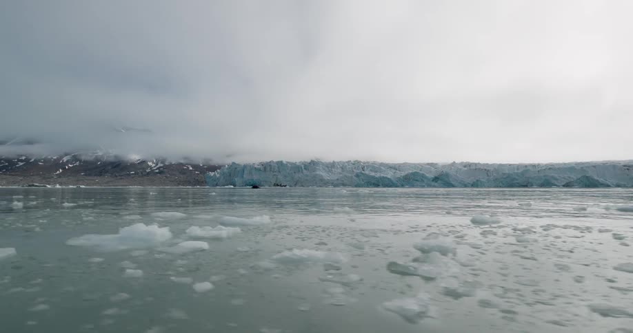 A vast glacier meeting the sea under a heavy overcast sky small chunks of ice float in the foreground of the cold grey water as low clouds cling to the surrounding mountain peaks