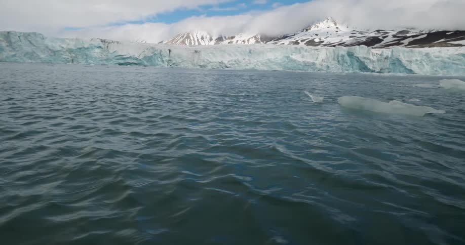 Dark rippling ocean water leads up to a massive glacier wall set against snowcapped mountains