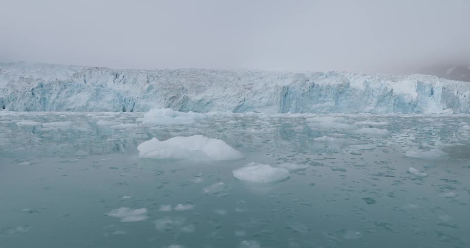 A massive arctic glacier meets calm icy waters dotted with floating ice chunks under a gray sky
