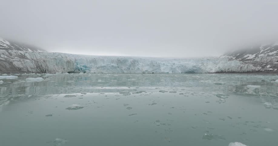 A massive white glacier spans the horizon under a heavy fogfilled skysmall chunks of ice drift across the calm dark water as the camera pans slowly to the left
