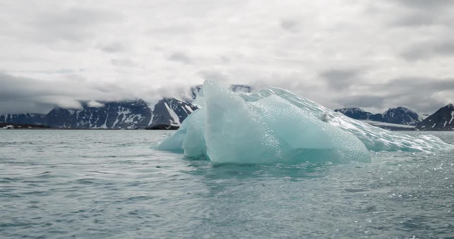 Bluish ice iceberg floating in cold water with distant mountains under a cloudy sky