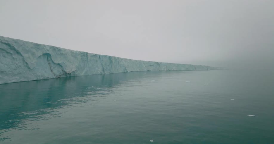 This video features a wide aerial shot of a massive glacier wall meeting the dark calm ocean under a heavy gray fog the camera slowly glides forward over the water revealing the immense scale and jagged texture of the ice shelf