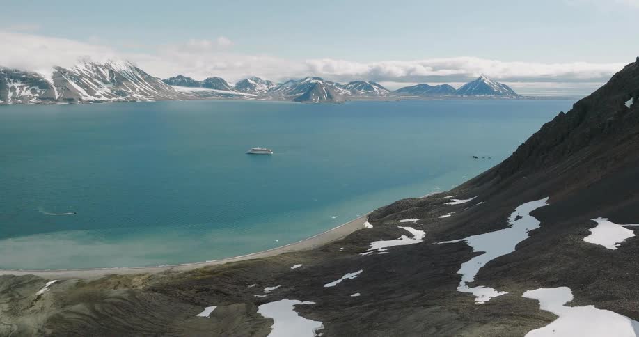 This video features a highangle drone shot of a cruise ship anchored in a calm arctic bay surrounded by snowdusted mountainsthe camera pans slowly across the rugged rocky coastline revealing small speedboats moving through the clear blue water nea