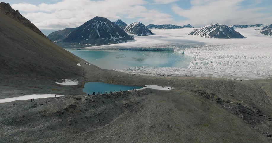 Extensive glacier reaching a glacial lake with snowcovered mountains in the background and a small group of people along the shore