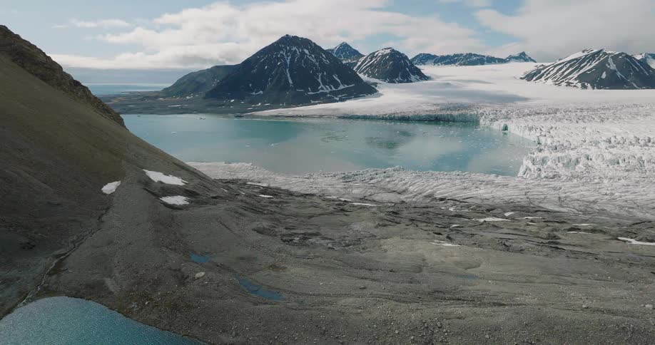 A group of hikers traverses a rocky ridge beside a turquoise pool before the view expands to reveal a massive glacier flowing between snowcapped peaks