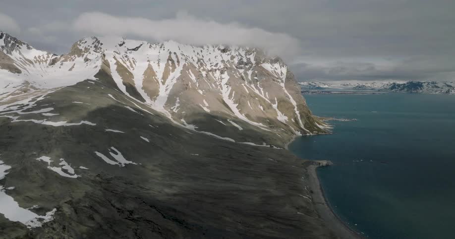 Coastal mountains with partially snowcovered slopes descending to a deepwater inlet under a cloudy sky