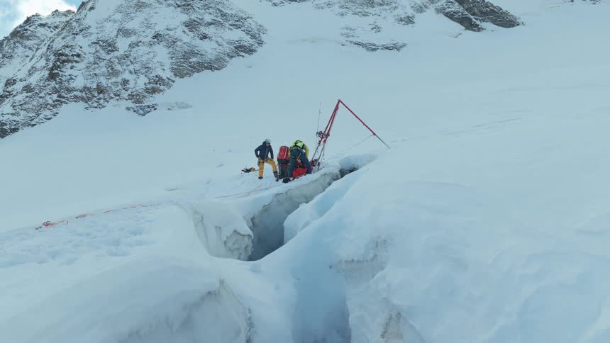 A team of people in bright winter gear works near a deep crevasse on a snow covered mountain using specialized equipment