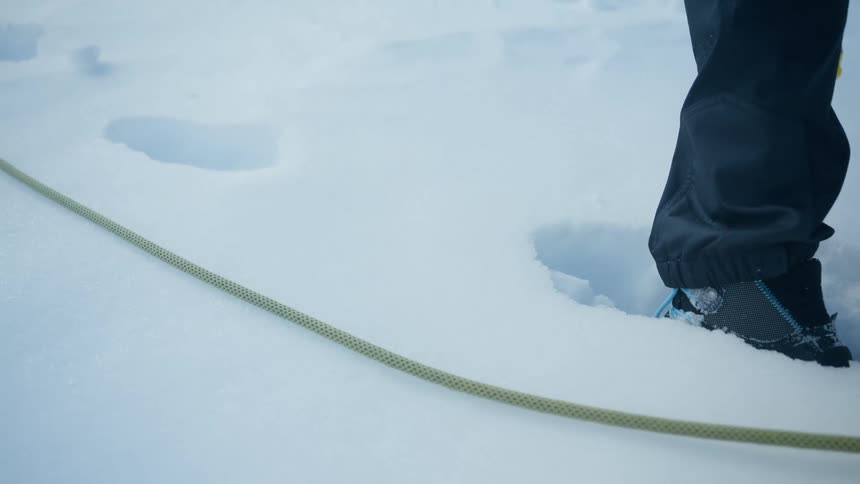 A close up view shows a person wearing crampons and dark pants walking through deep snow next to a yellow climbing rope