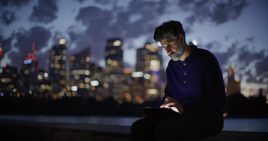 A man with a saltandpepper beard sits on a ledge at night focused on using a tablet and stylusbehind him a sprawling city skyline glows with golden lights against a deep blue twilight sky