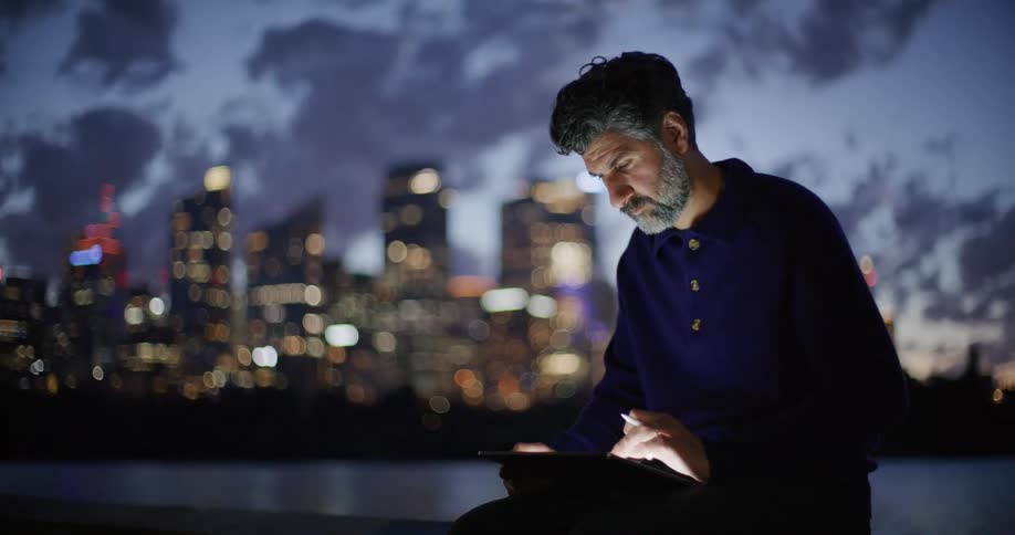 Man uses a tablet at night with an illuminated city skyline in the background