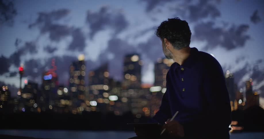 Man sitting along the coast working on a tablet computer with city buildings in the distance
