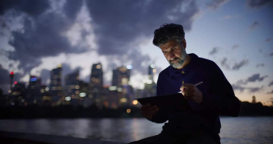Man sitting along the coast working on a tablet computer with city buildings in the distance