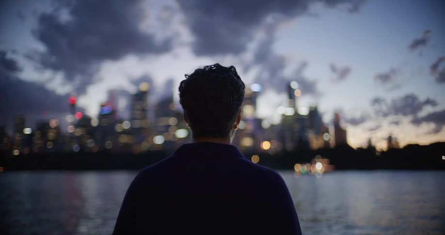 Person seen from behind observing a lit city across a body of water at dusk