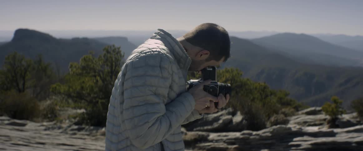 A person aims a camera while standing on a rocky ledge