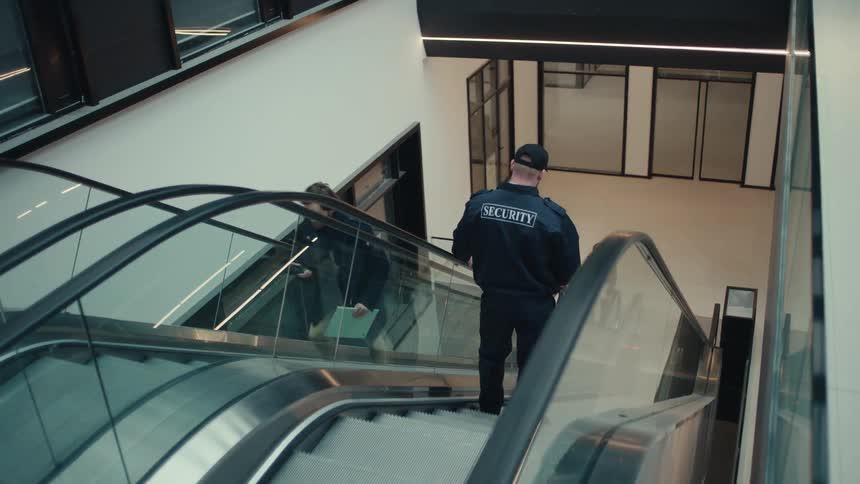 A security guard rides down an escalator inside a modern building while monitoring the area