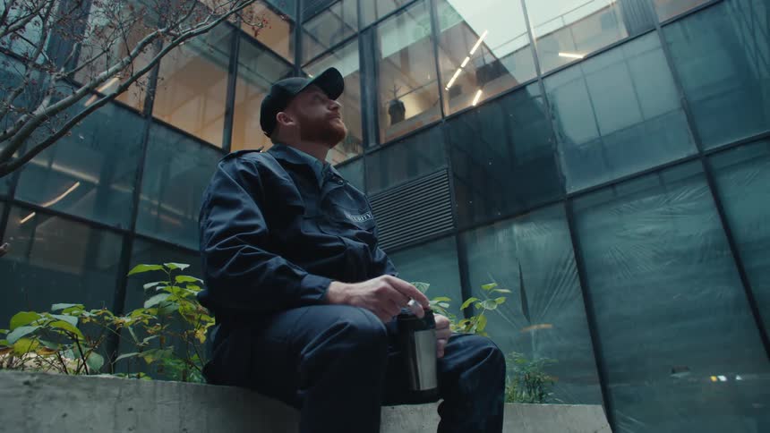 A security guard smokes and drinks from a travel mug while taking a break outside a modern glass building