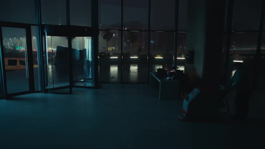 Dark office lobby at night a security guard sits at a desk while a large revolving door remains still on the leftas the scene progresses a cleaning person enters from the right pushing a floor scrubbing machine across the polished floor toward the e
