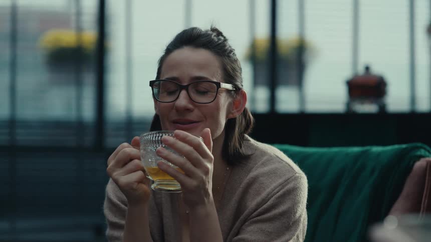A woman wearing glasses sits on a couch holding a glass of tea and engaging in a cheerful conversation with someone offcamera she takes a slow sip from her cup with her eyes closed in a moment of relaxation before continuing her discussion with a smile