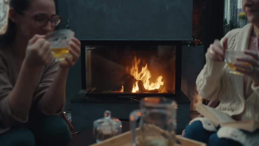 Mother and daughter sit indoors enjoying warm drinks while chatting together by a fireplace