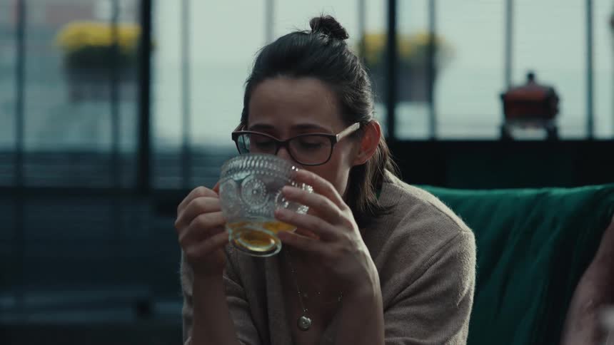 A middle aged woman with her hair in a bun and wearing glasses sits in a modern room smiling and talking while holding a decorative glass cup of tea