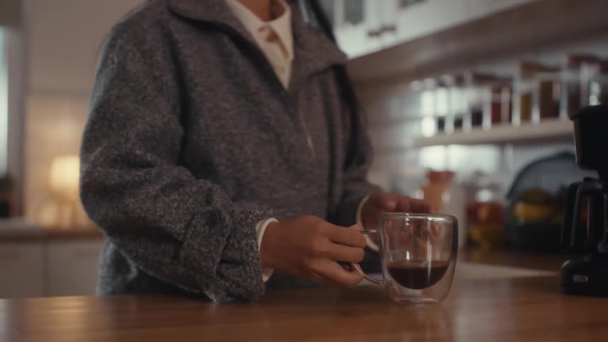 Woman stands in the kitchen pouring coffee into a glass cup and taking a drink