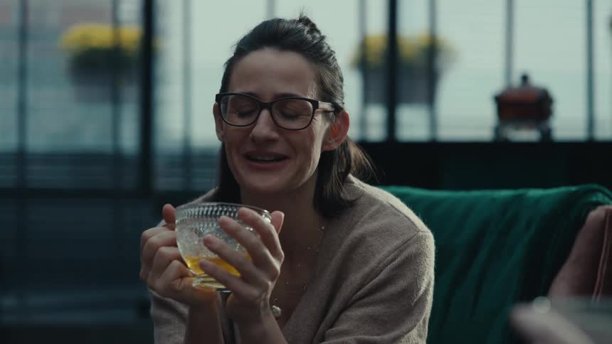 A young woman sits in the living room wearing glasses sipping a cup of tea while talking with another person