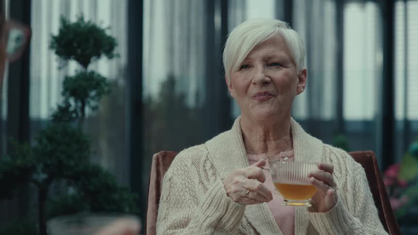 A smiling older woman with short white hair drinks tea while chatting with a companion