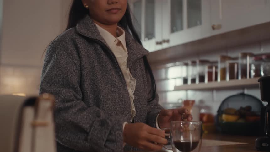 A young woman pours coffee into a glass cup and takes a sip in her kitchen at midday while wearing a winter jacket