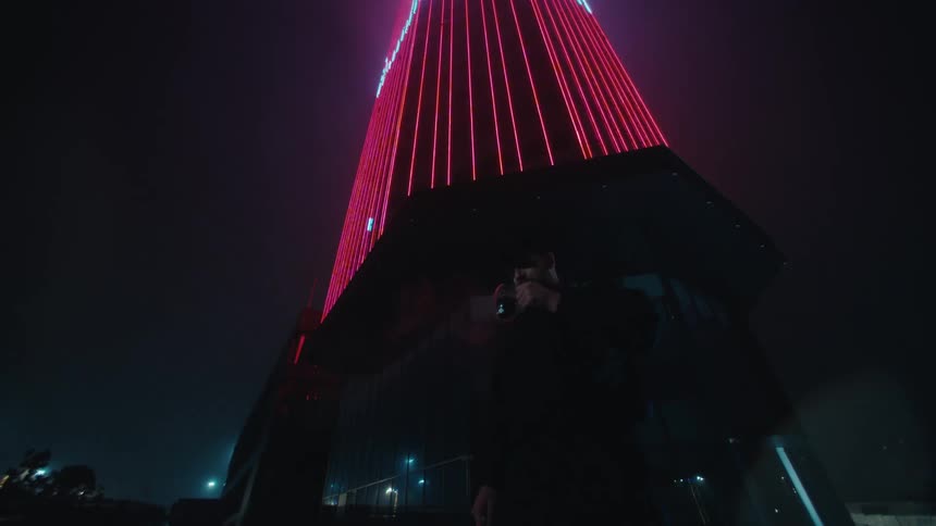 A young man stands outside a building at night smoking a cigarette while the side of a nearby highrise glows red and cars pass by on the street