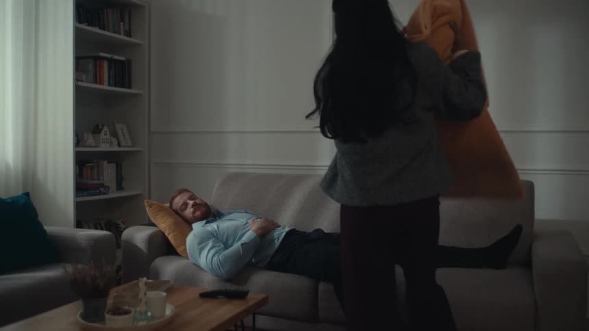 A woman gently removes the remote from a sleeping man on the sofa and covers him with a blanket