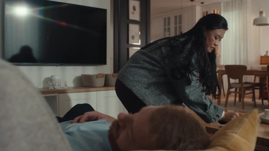 Woman covers a man lying down with a blanket in a living room with a kitchen in the background