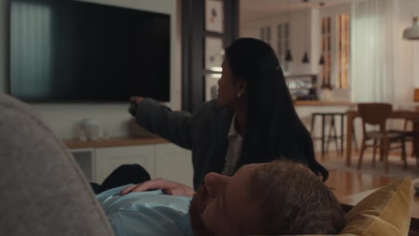 A young woman in a grey jacket walks from a kitchen into a living room where a man is asleep on a couch she picks up a remote to turn off the television and then covers the sleeping man with a tan blanket