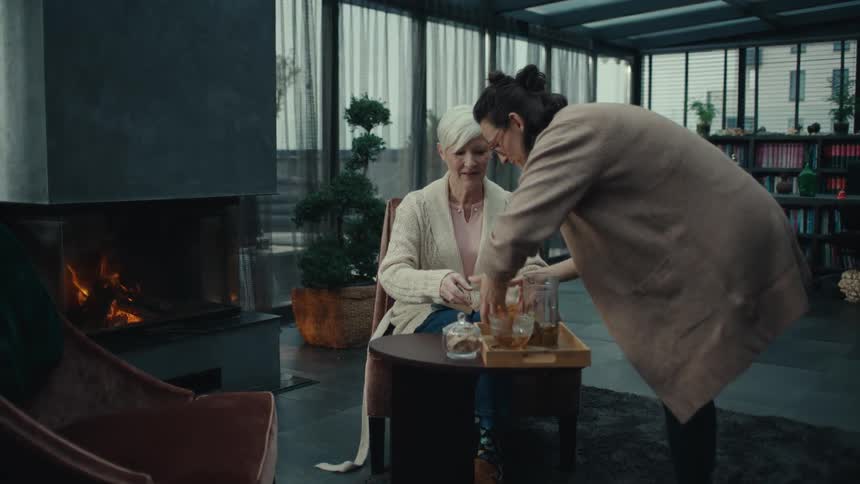 A younger woman brings a tray of tea to an older woman in a sunroom and sits down to join her