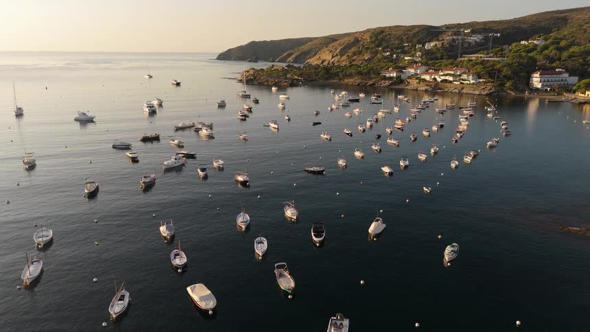 An aerial view shows many small white boats and a few red rafts floating on calm dark water in a coastal bay